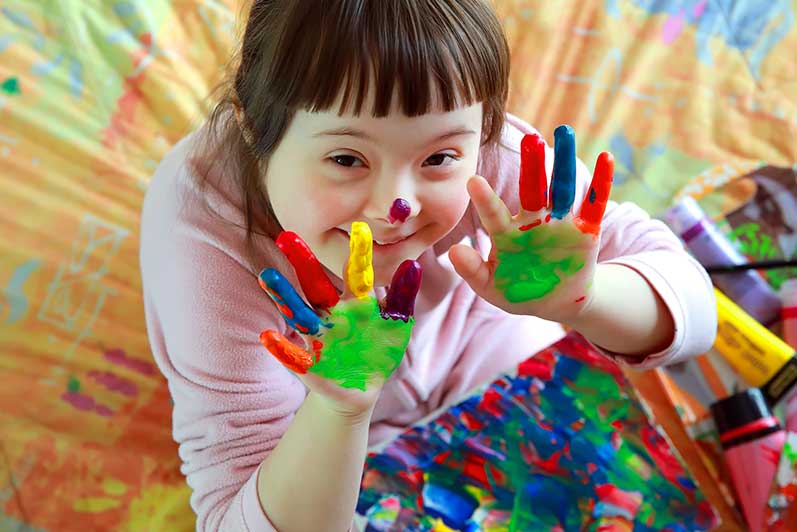 Photo of girl with down syndrome smiling while showing her hands covered in colorful paint